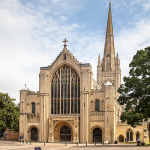 Norwich Cathedral sits next to a large tree. The sky is blue and cloudy.