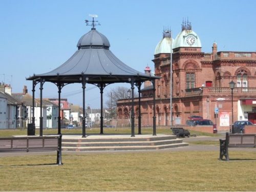 Wide angle shot of Gorleston-on-Sea pavillion. It is surrounded by green grass with a red brick bulding behind it.