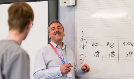 David Stowell, a white man with grey hair and a grey moustache, stands in front of a whiteboard. The white board has music annotation on it. He is smiling at a student, who has red hair and glasses.