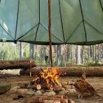 A small campfire burns on the ground inside a tent-like shelter made of green fabric, surrounded by logs in a forest setting.