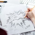 A person holding a pencil drawing the word 'BOOM' in bold letters surrounded by explosive lines on a piece of sketch paper,